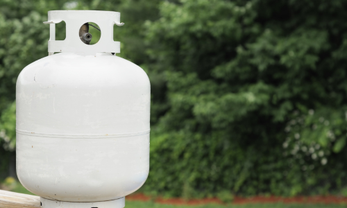 Close-up of a propane tank with a brass valve in front of an open outdoor grill. The scene conveys readiness for a barbecue, set in a green backyard.